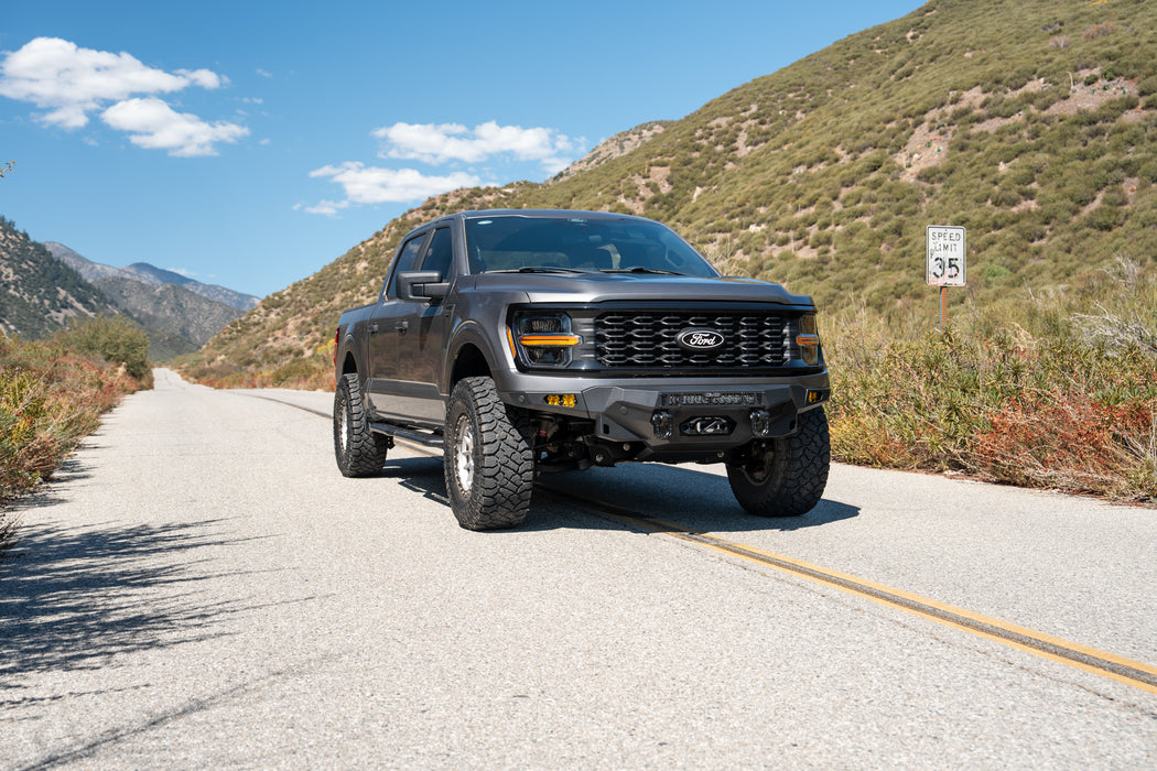 A dark gray 2024+ Ford F-150 with DV8 Spec Series front bumper, driving on a paved mountain road under a blue sky.