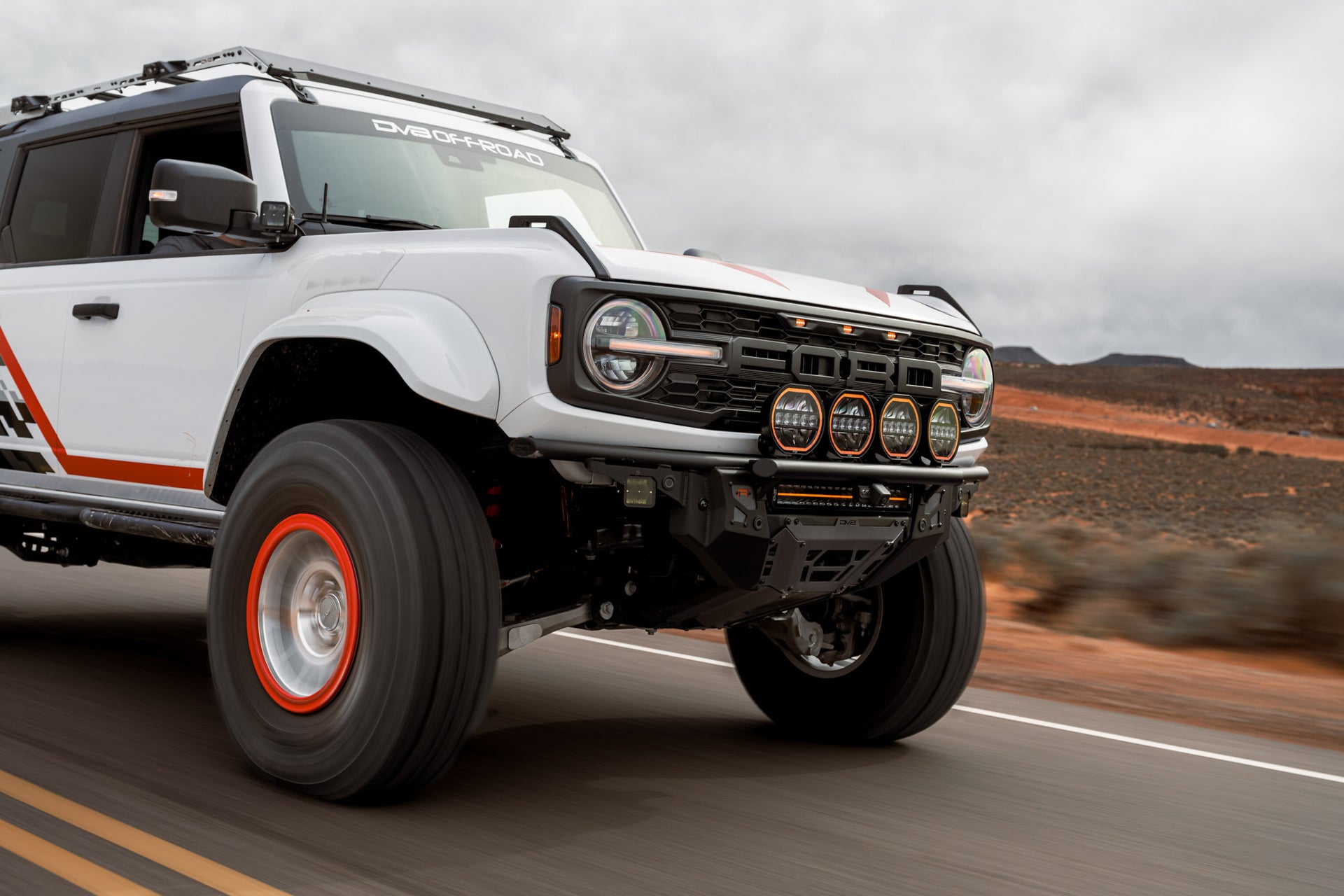 White Ford Bronco Raptor with Tube Front Bumper driving on a road with a desert landscape in the background.
