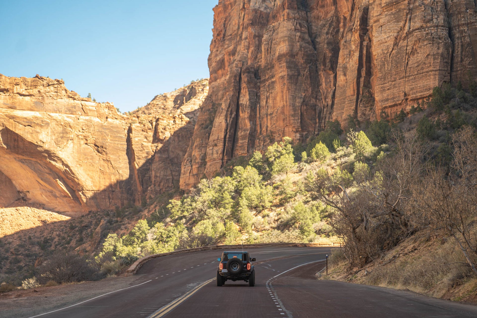 A Bronco drives along a winding road between towering orange-brown sandstone cliffs and green shrubs under a clear blue sky.
